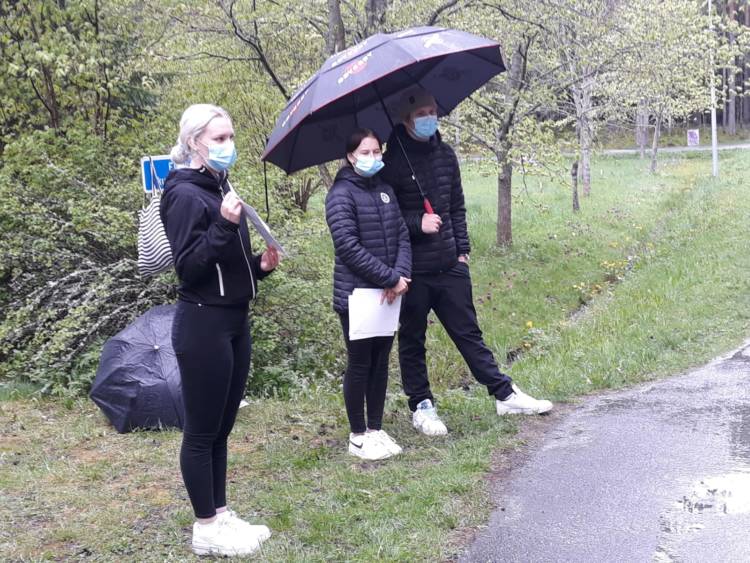 Kolme tummiin pukeutunutta henkilöä maskit kasvoillaan ulkona. Kaksi seisoo sivummassa sateenvarjon alla ja yksi esittelee valkoista paperialappua.