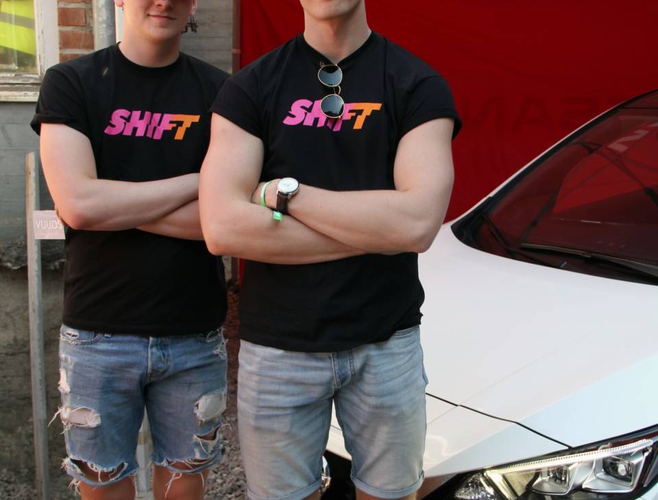 Kaksi henkilöä Shift-t-paidoissa henkilöauton edessä.