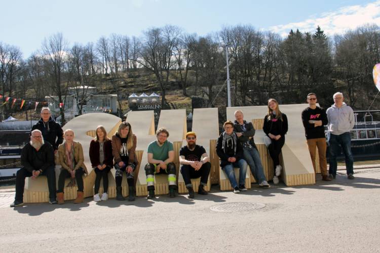 Henkilöt istuvat auringon paisteisessa kelissä vanerista valmistettujen SHIFT-kirjaimien päällä.