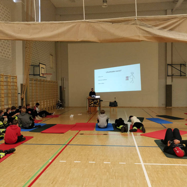 Tubettaja HardKokki pitää luentoa opiskelijoille liikuntasalissa. Opiskelijat kuuntelevat lattialla jumppamattojen päällä istuen ja leväten.