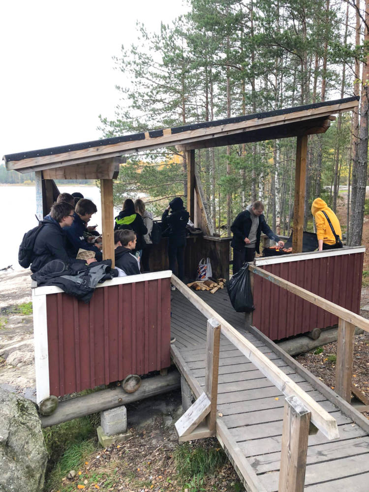 Opiskelijoita katoksessa. Yksi paistaa makkaraa, loput istuvat tai seisovat katoksen reunustoilla.