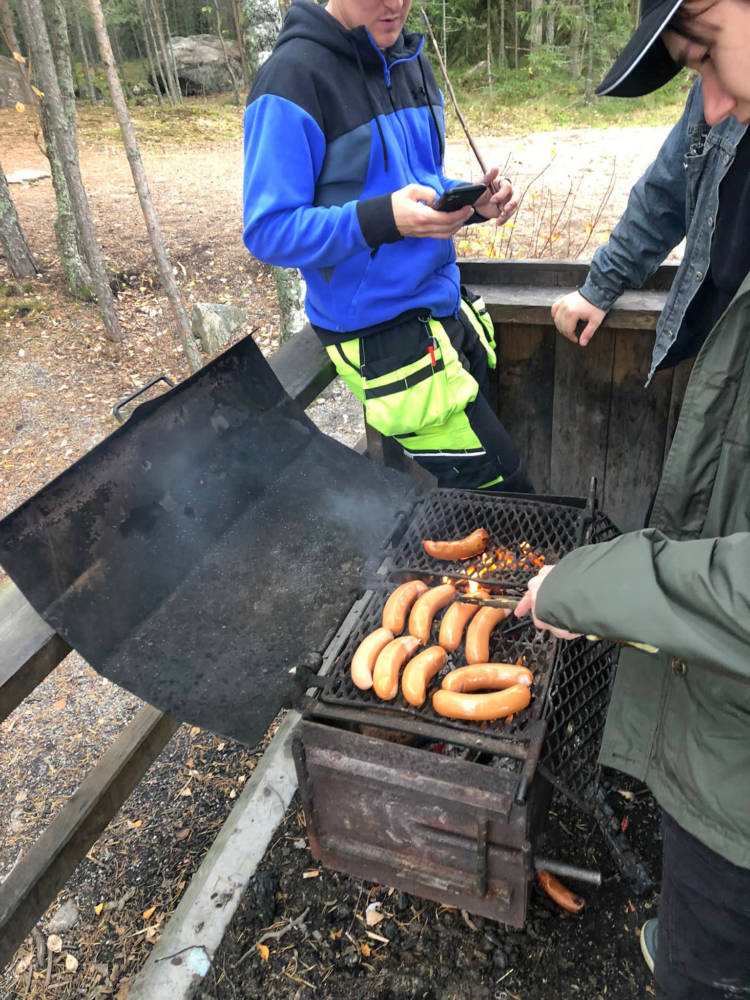 Opiskelija paistaa makkaraa. Kaksi muuta opiskelijaa seisovat vieressä.