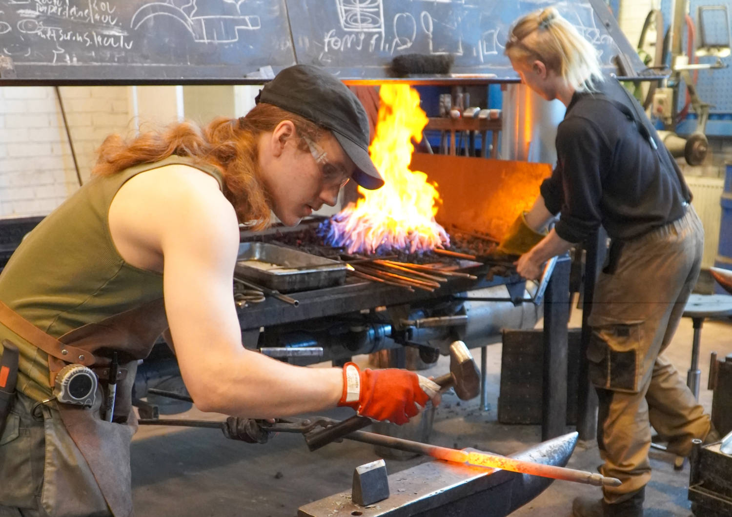 Kaksi henkilöä metalliseppien työsalissa. Toinen kuumentaa metallia tahkolla ja toinen takoo alasimella.