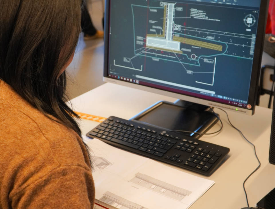Suunnitteluassistenttiopiskelija tietokoneella. Samalla hän mittaa paperilla olevaa rakennuksen teknistä kuvaa.