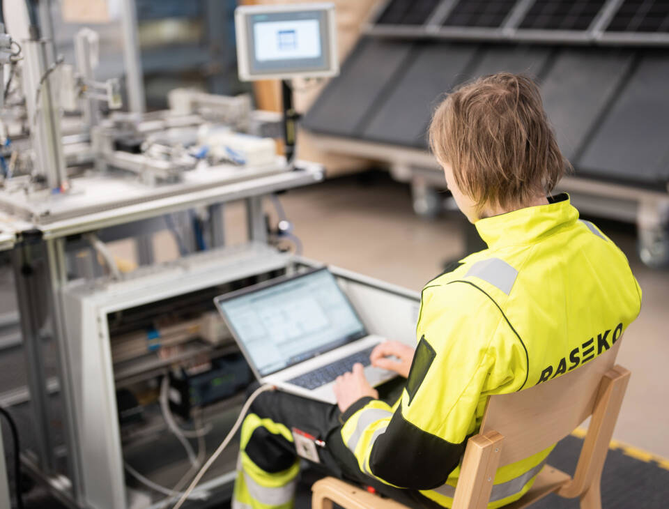 Rasekon työvaatteisiin pukeutunut henkilö tietokone sylissään automaatiolinjan ääressä.