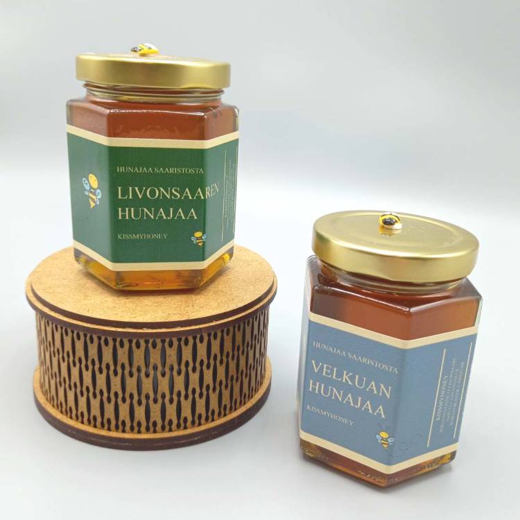 Kaksi lasista hunajapurkkia, joista toinen lieriön muotoisella puisella korokkeella. Toisessa purkissa vihreä etiketti, jossa lukee "Livonsaaren hunajaa," Toisessa sininen etiketti tekstillä "Velkuan hunajaa."