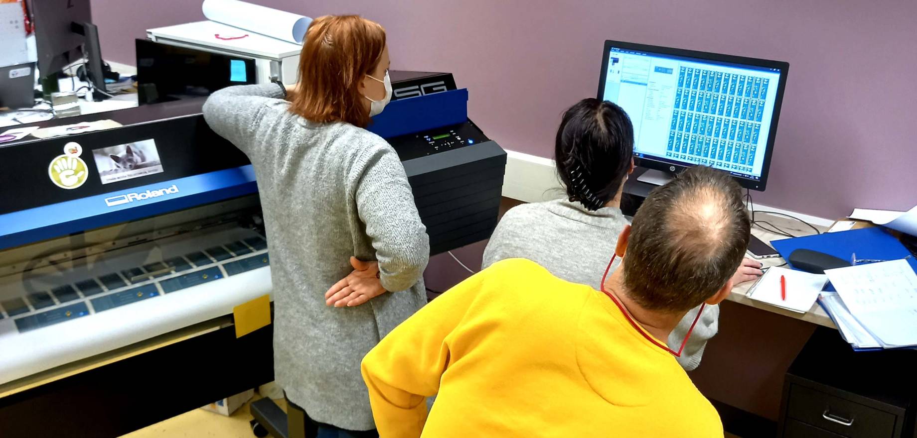 three people next to a large scale printer, looking at computer screen with graphic design elements, with a backdrop of purple wall.