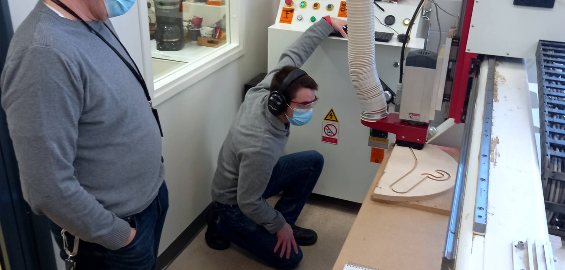Two people in grey shirts next to a CNC machine. One of them is standing, the other one kneeling. Both are wearing hearing protection and protective eyewear and facemasks