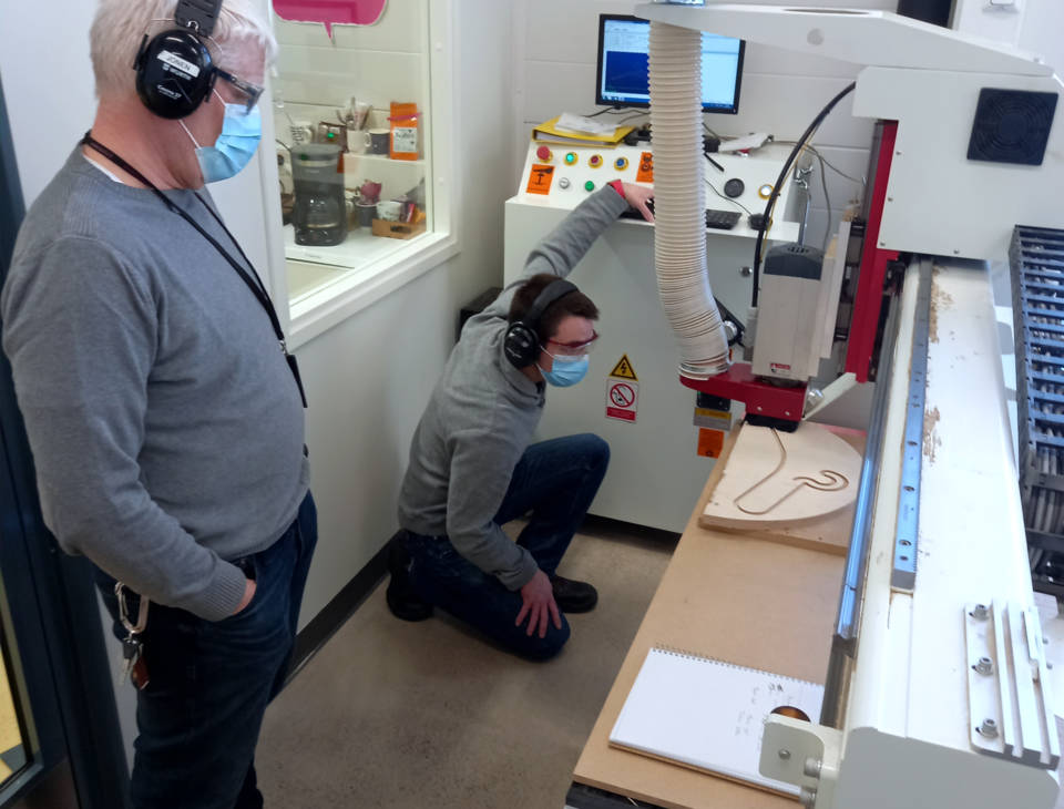 Two people in grey shirts next to a CNC machine. One of them is standing, the other one kneeling. Both are wearing hearing protection and protective eyewear and facemasks