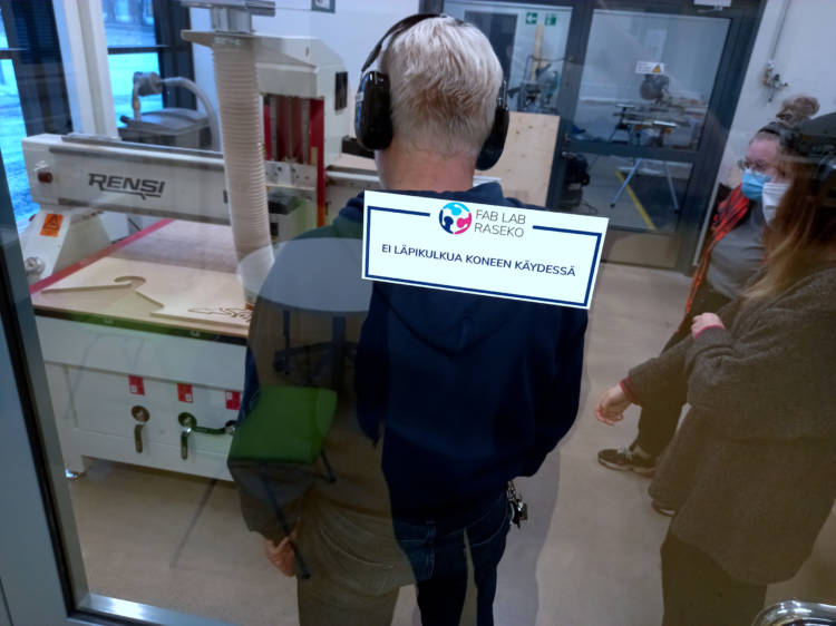 View through a glass door. The door has a sticker with Fab Lab Raseko logo and reads in Finnish "No entry while machine in use." Behind the door stands a person in dark blue shirt, wearing hearing protection. Behind them is a CNC machine.