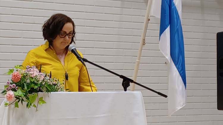 Henkilö puhuu mikrofoniin pöydän takana. Pöydällä on kukkakimppu ja vieressä lattialla suomenlippu.
