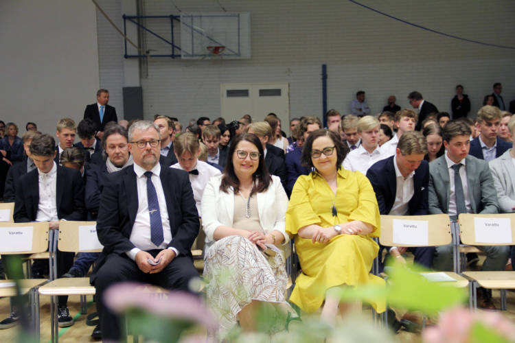 Paljon yleisöä istumassa juhlasalissa. Edustalla kolme henkilöä istuu tyhjällä penkkirivillä.