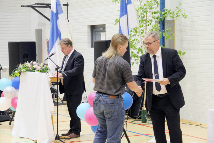 Kaksi henkilöä kättelevät ja toisella on todistus kädessään. Kolmas henkilö seisoo taustalla puhujanpöydän takana.