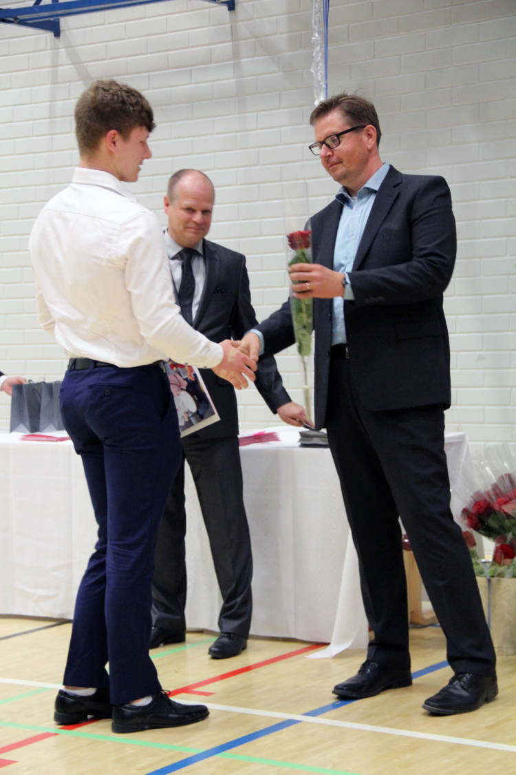 Kaksi henkilöä kättelee ja toinen on ojentamassa toiselle ruusua. Taustalla seisoo kolmas henkilö.