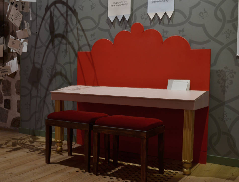 Museotilassa on punaisen sävyinen pöytä, jonka takaseinän kuvio on aaltoileva ja jalat kultaiset sekä kaksi jakkaraa.