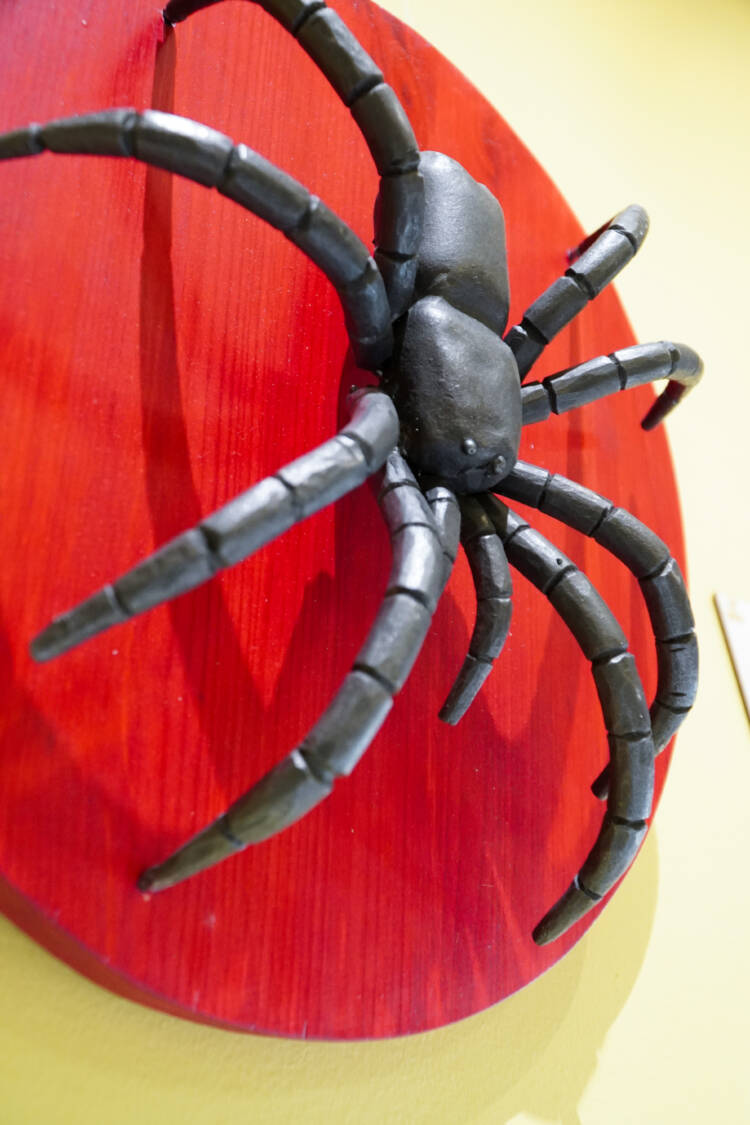 Metallista taottu hämähäkki punaiseksi maalatun, ympyränmuotoisen, puusta tehdyn levyn päällä.