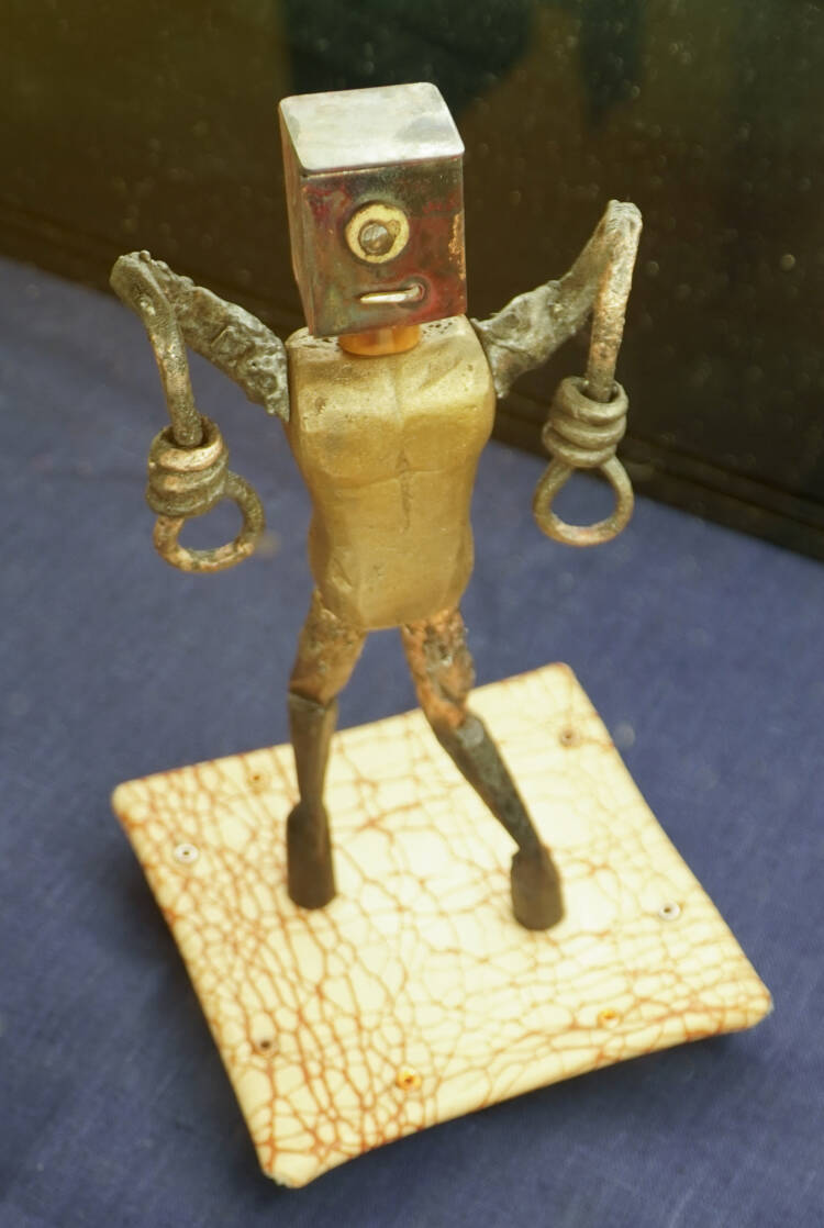Robotin ja ihmishahmon muotoinen metallinen teos nahkaisen alustan päällä. Hahmon pää on kuution muotoinen ja sen kädet roikkuvat. Käsien päissä on silmukat. Vieressä pöydällä on kyltti, jossa lukee Moxie Contin, Viivi Björklund.
