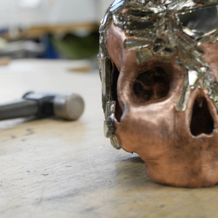 Metallinen pääkalloveistos puisella tasolla, takana utuisena vasara.