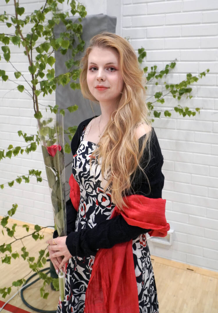 Nuori pitkätukkainen juhlapukuinen henkilö ruusun kanssa koivujen edessä.