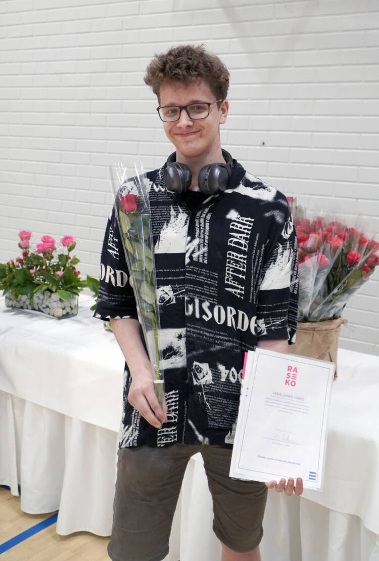 Nuori hymyilevä henkilö stipendin ja ruusun kanssa valkoisen pöydän edessä.