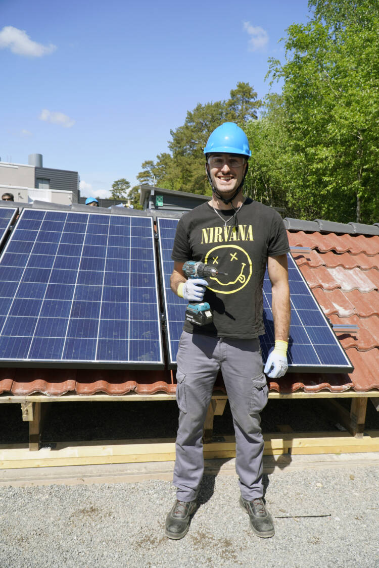 A helmeted man with a drill in his hand in front of a solar panel.