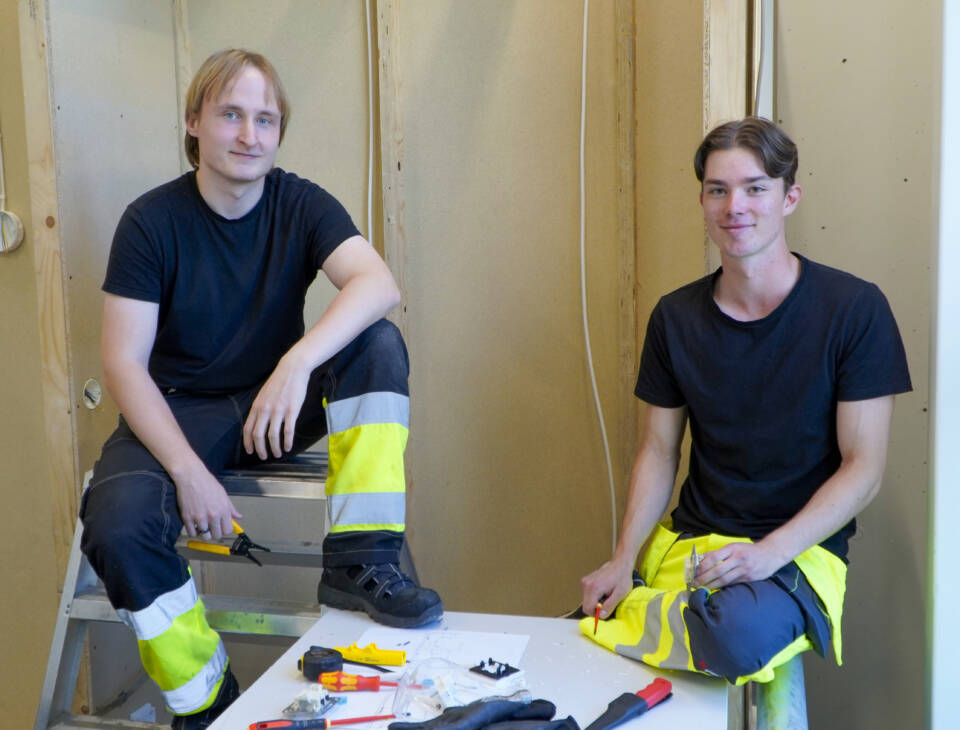 Kaksi henkilöä työvaatteissa istuvat vierekkäin sähköalan työsalissa.