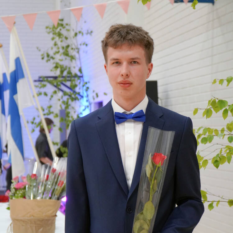 Tummansiniseen pukuun pukeutunut henkilö seisoo juhlatilassa ruusu kädessään.