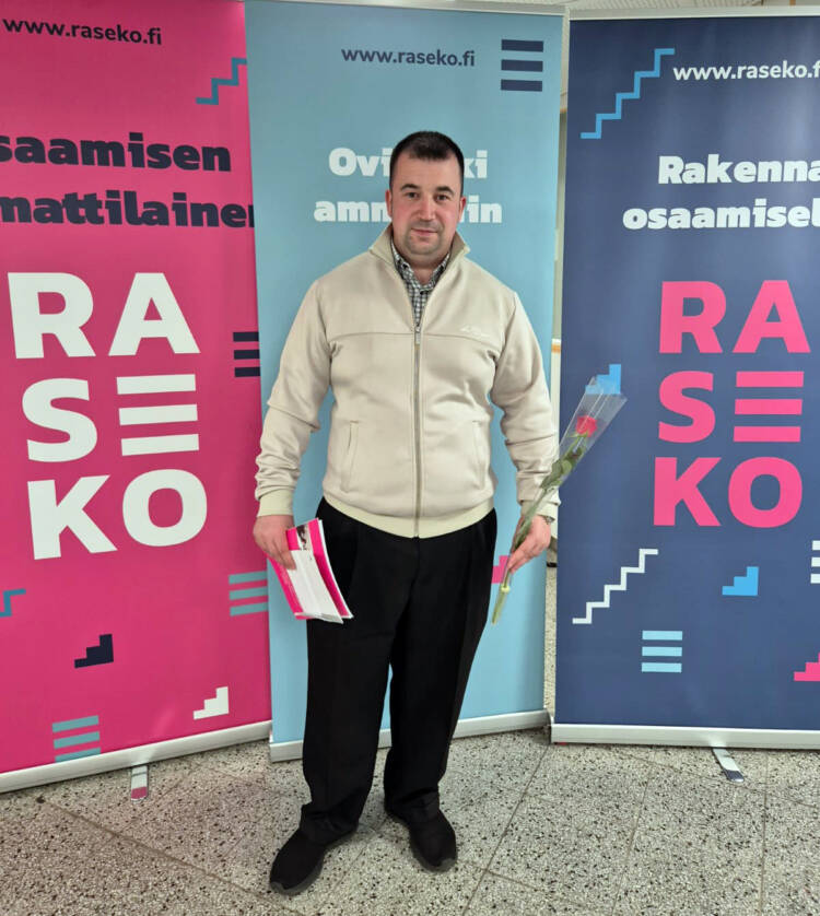 Henkilö seisoo kolmen Rasekon roll-upin edessä ruusu ja lehtinen kädessään.