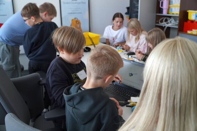 Lapsia työskentelmässä LEGO robottien kanssa pitkän pöydän ääressä, nuoria ohjaamassa heitä.