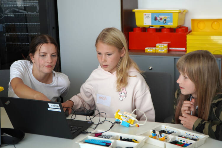 Kaksi lasta kokoamassa LEGO robottia, nuori ohjaamassa heitä ja osoittaa tietokonetta.